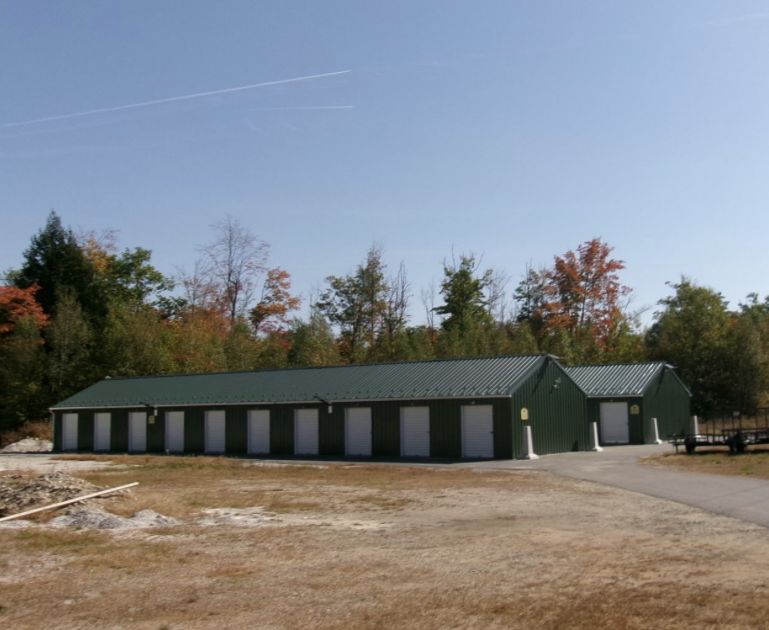 PB&J Self Storage in Limerick, Maine: a long green steel storage building with white roll-up doors and a paved drive, with fall foliage behind